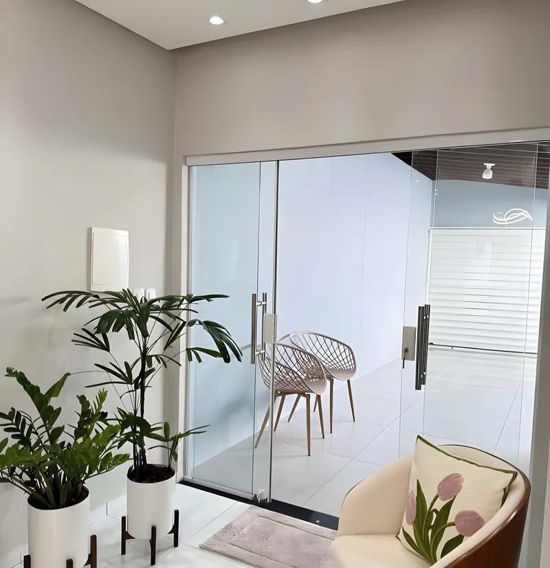 Porta de vidro temperado de correr com puxadores em inox, ao lado de poltrona e plantas